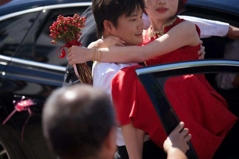 新郎从黑色轿车后座抱出穿红裙的新娘，新娘手持红色果实花束，前景是一位长辈的后脑勺。